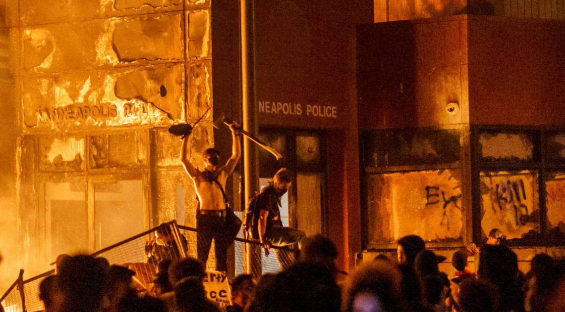 Chamas de um incêndio próximo iluminam os manifestantes em pé em uma barricada em frente a delegacia de polícia em 28 de maio de 2020 em Minneapolis, Minnesota