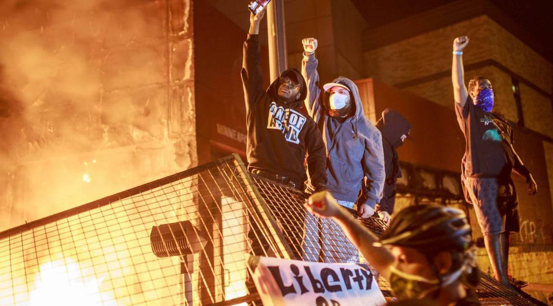 Manifestantes erguem os punhos enquanto as chamas se erguem atrás deles na frente da delegacia de polícia 