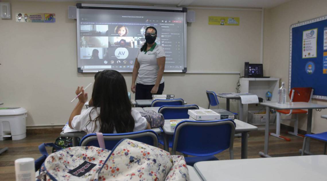 Professora d&aacute; aula para duas crian&ccedil;as presencialmente, enquanto outras acompanham por v&iacute;deo