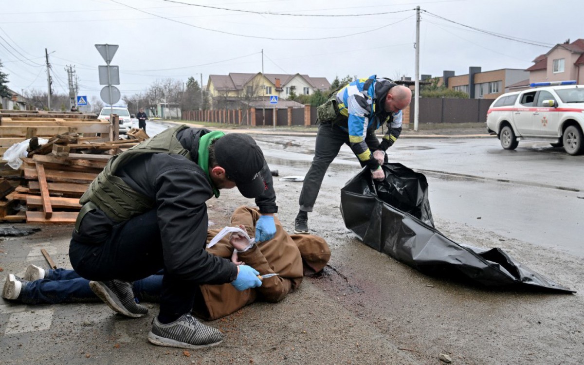Trabalhador corta a corda do cadáver de um homem com as mãos amarradas atrás das costas enquanto um colega prepara um saco plástico para carregá-lo para um carro na cidade de Bucha, na Ucrânia - SERGEI SUPINSKY/AFP