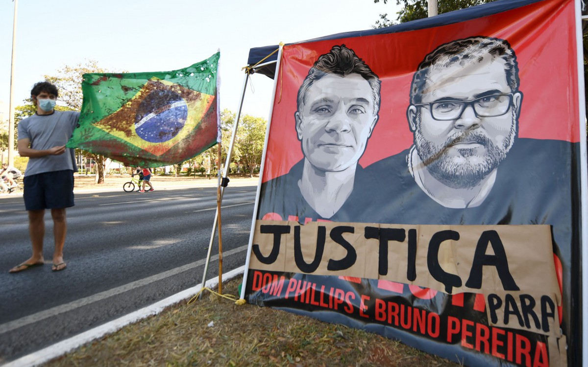 A man holds a Brazilian flag tinted in red representing blood next to a banner in demand of justice for the kinning of British journalist Dom Phillips and Brazilian indigenist Bruno Pereira, during a protest in Brasilia, on June 19, 2022. Phillips and Pereira were killed in the Amazon by illegal fishermen and their bodies buried in the middle of the forest in the valley of Javari river.
EVARISTO SA / AFP


 - EVARISTO SA / AFP