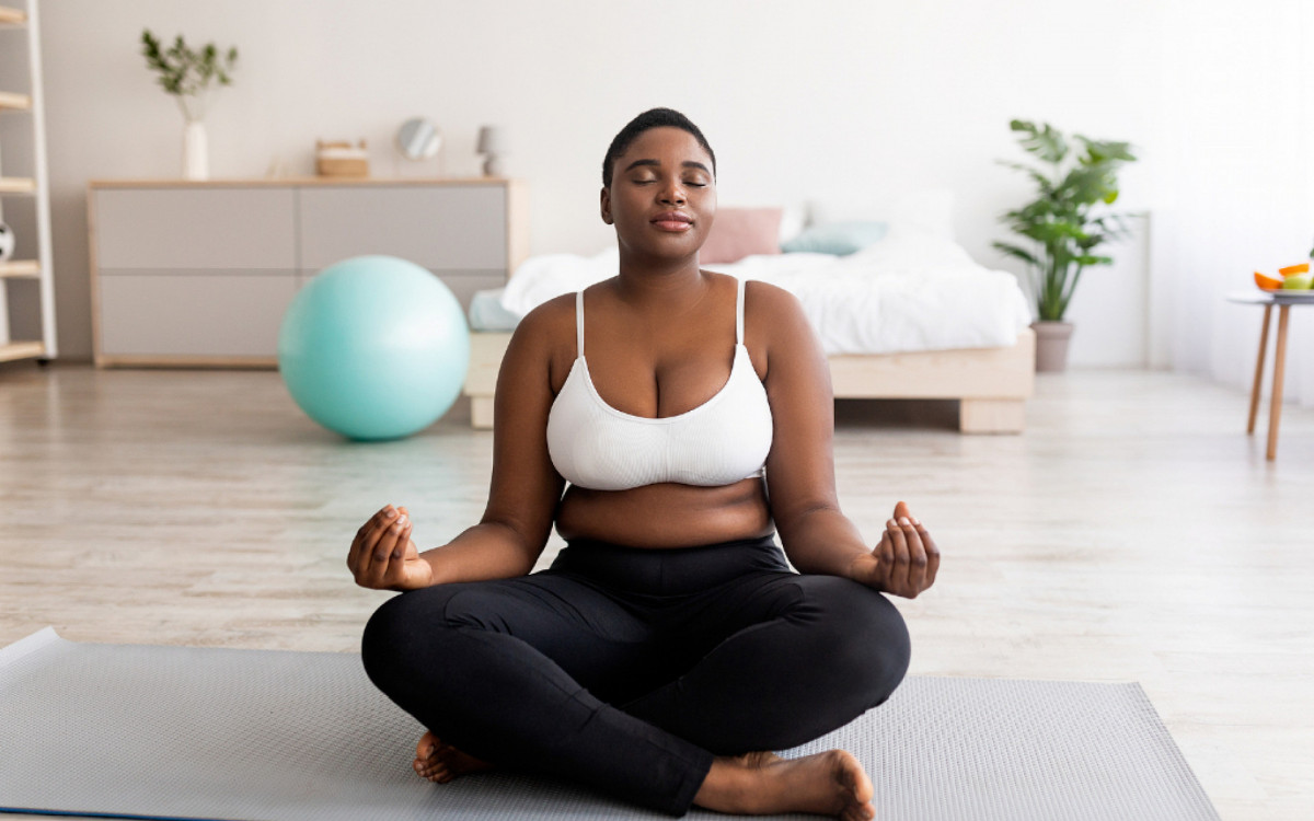 Plus Size Black Woman Sitting In Lotus Pose, Meditating With Closed Eyes, Practicing Yoga At Home