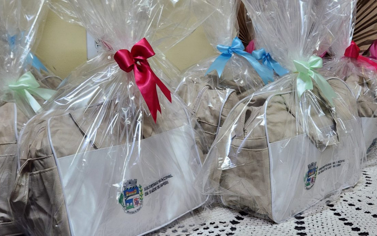 O kit enxoval entregue é composto por: bolsa, trocador, macacão, toalha de banho com capuz, manta, meias, touca, luva e banheira