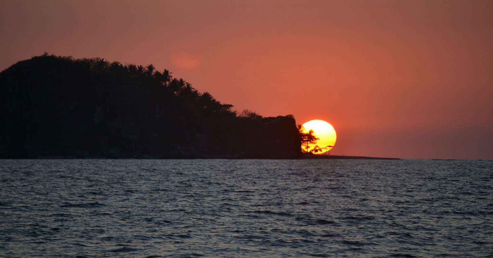 A beautiful wide silhouette shot of an islet covered with trees on by the sea under sky during sunset