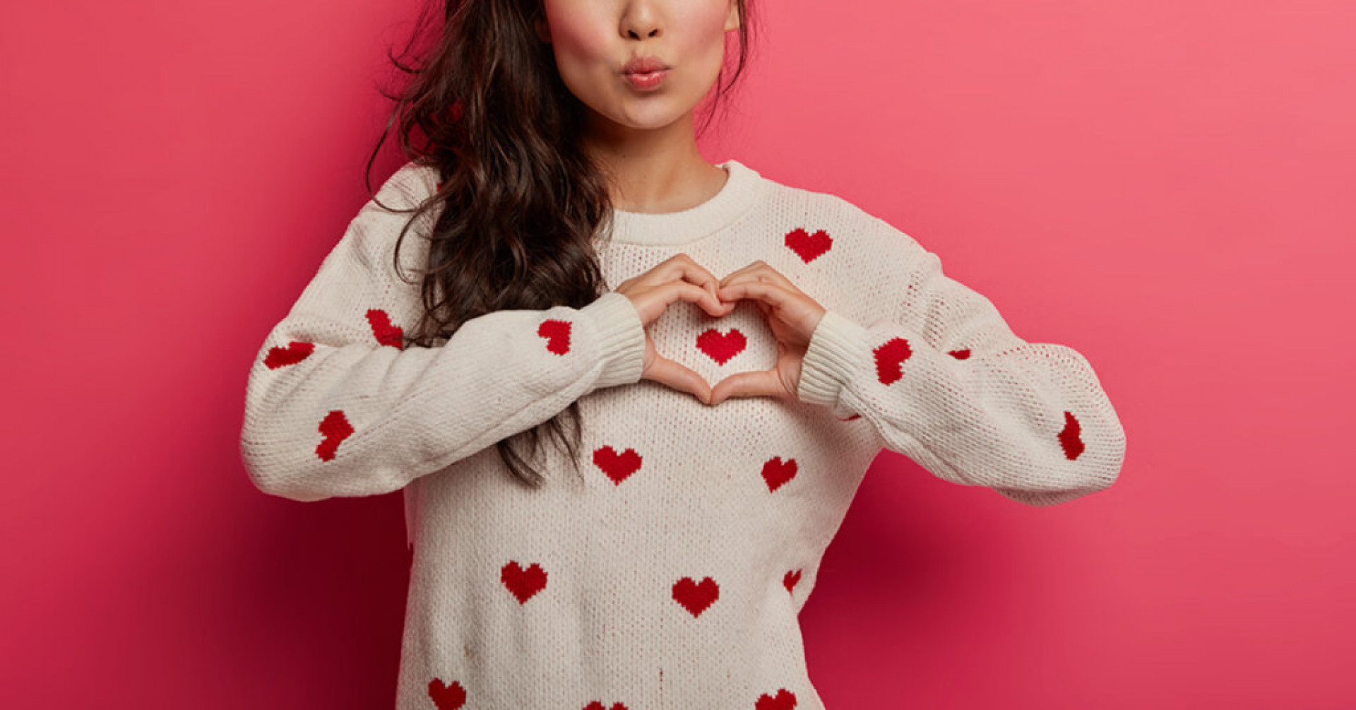 Charming woman with folded lips shows heart gesture over chest, expresses heartwarming feelings, sympathy and tender, dressed in casual sweater, models against pink background. Love and admiration