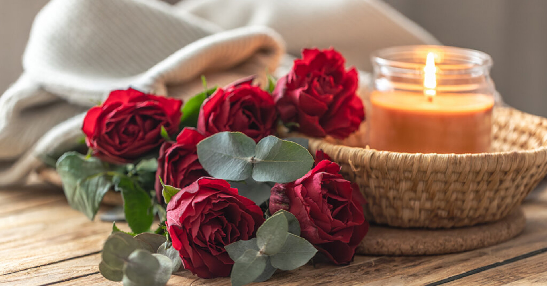 Lit candle in a glass jar in a woven tray with red roses and eucalyptus on warm wood. Soft daylight builds hygge cozycore mood for slowliving selfcare moments.